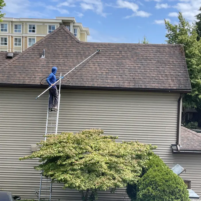 Technician on a ladder cleaning a multi-unit roof with an extension pole, apartment building behind, Surrey BC